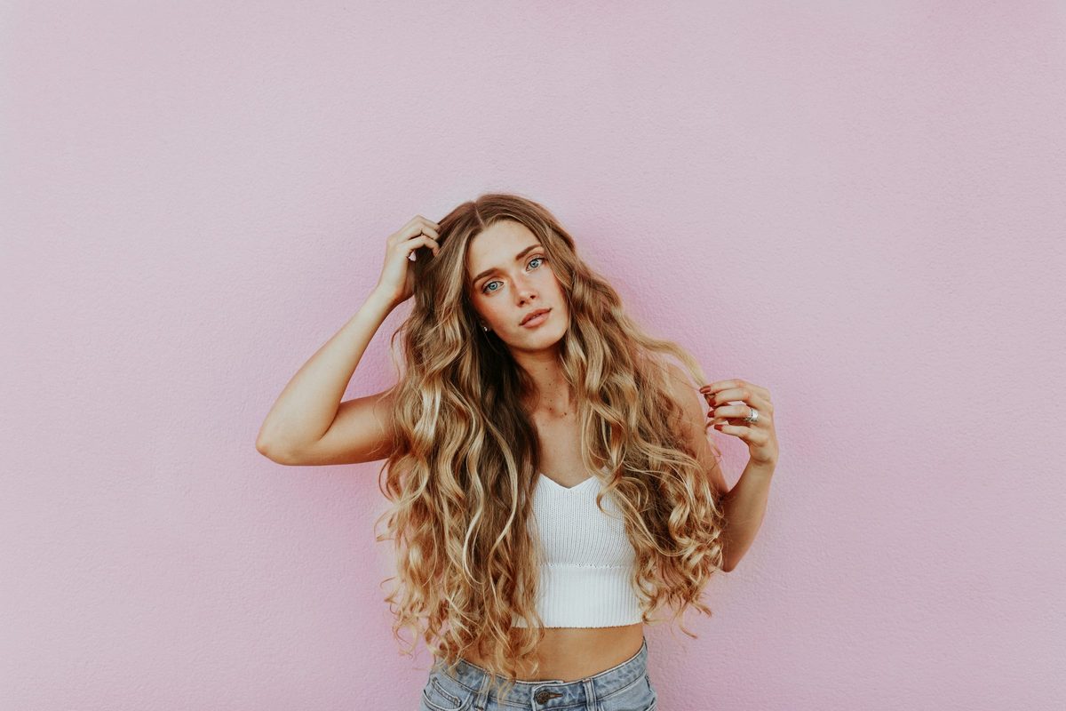 A woman with long, softly waved honey blonde hair, standing against a pink wall