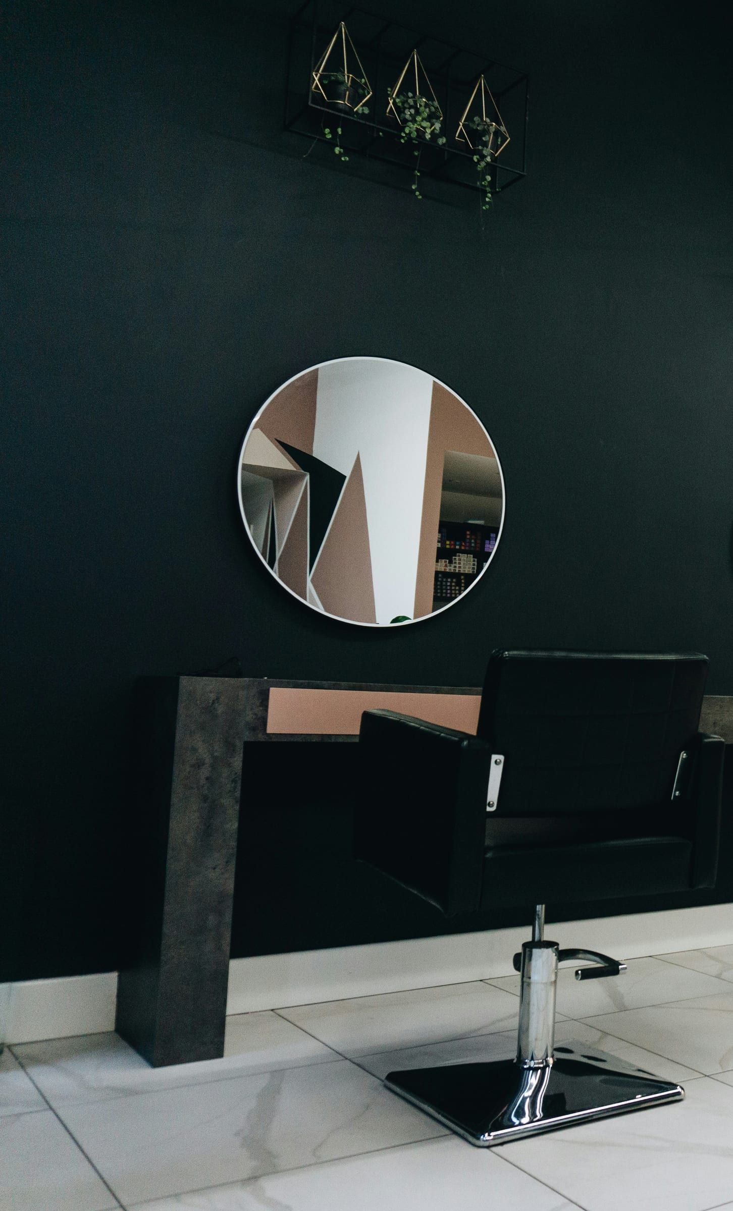 A round mirror and a black styling chair against a dark wall, with trailing plants above