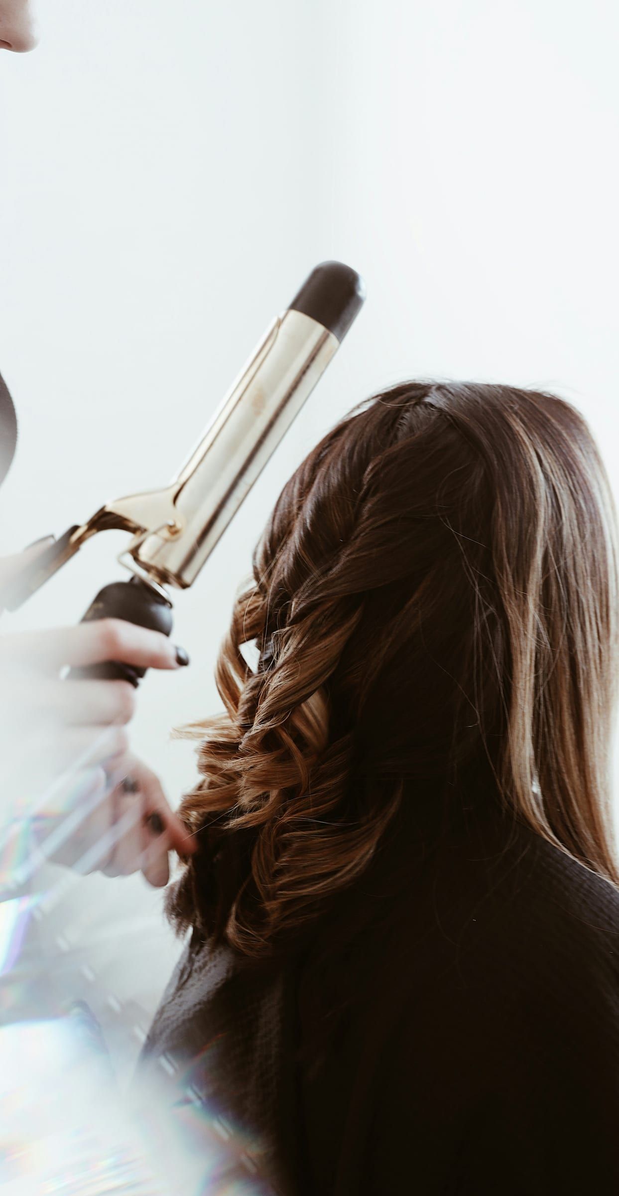 A stylist curling a client's brunette balayage with gold tongs